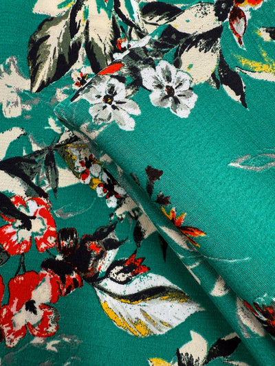 Close-up of Printed Viscose - Tropic Bloom (140cm) from Super Cheap Fabrics, featuring a vibrant green base with red, white, yellow, and black flowers plus green and cream leaves in a layered floral pattern—sourced from Prato, Italy.