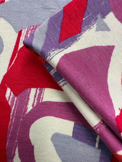 A close-up of Super Cheap Fabrics' Viscose Knit - Abstract Striped Geo (150cm) shows bold brushstroke patterns and overlapping colors in red, purple, gray, and off-white on soft, stretch fabric.