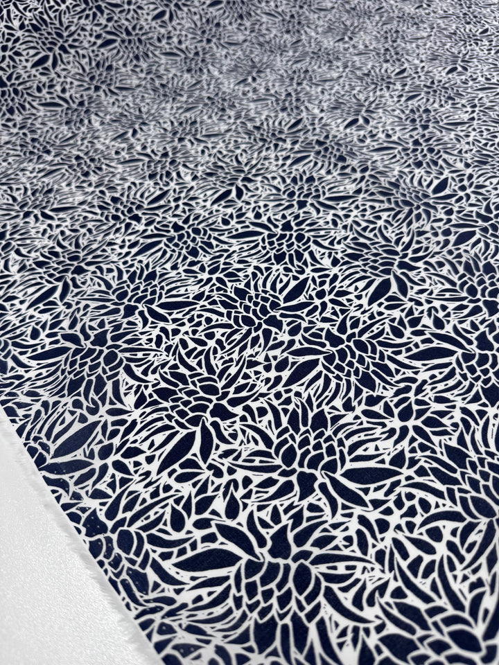 A close-up of Super Cheap Fabrics' Cotton Voile - Lotus - 147cm shows a textured surface with interwoven white floral and leaf designs on a dark blue background, creating a lace-like effect perfect for warm weather clothing.