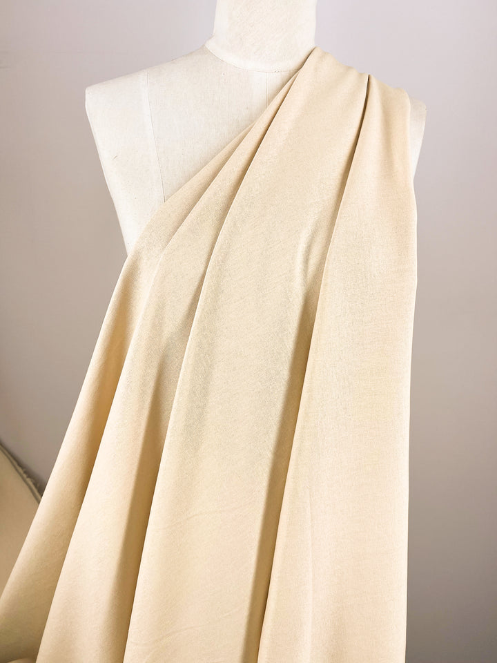 A mannequin is adorned with Premium Linen Cotton in Shortbread, a product of Super Cheap Fabrics. The elegant fabric creates graceful folds as it drapes over one shoulder and cascades down the front, offering a soft and classic look against a neutral background.