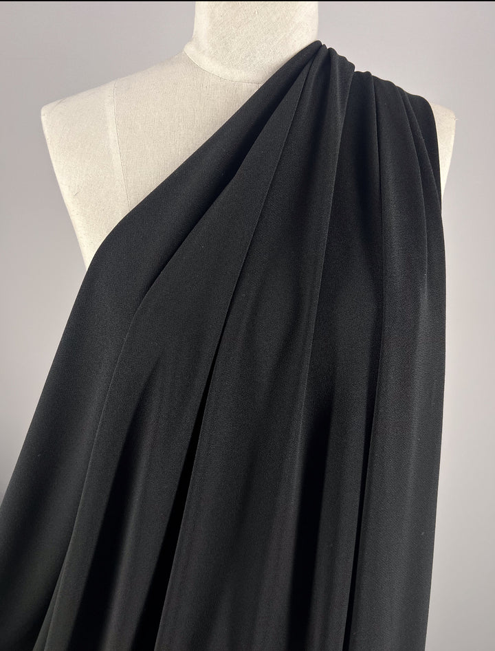 A close-up of a mannequin showcases "Plain Lycra - Black - 165cm" by Super Cheap Fabrics, draped in pleats over one shoulder like a toga. A plain background highlights its modern activewear-inspired style.