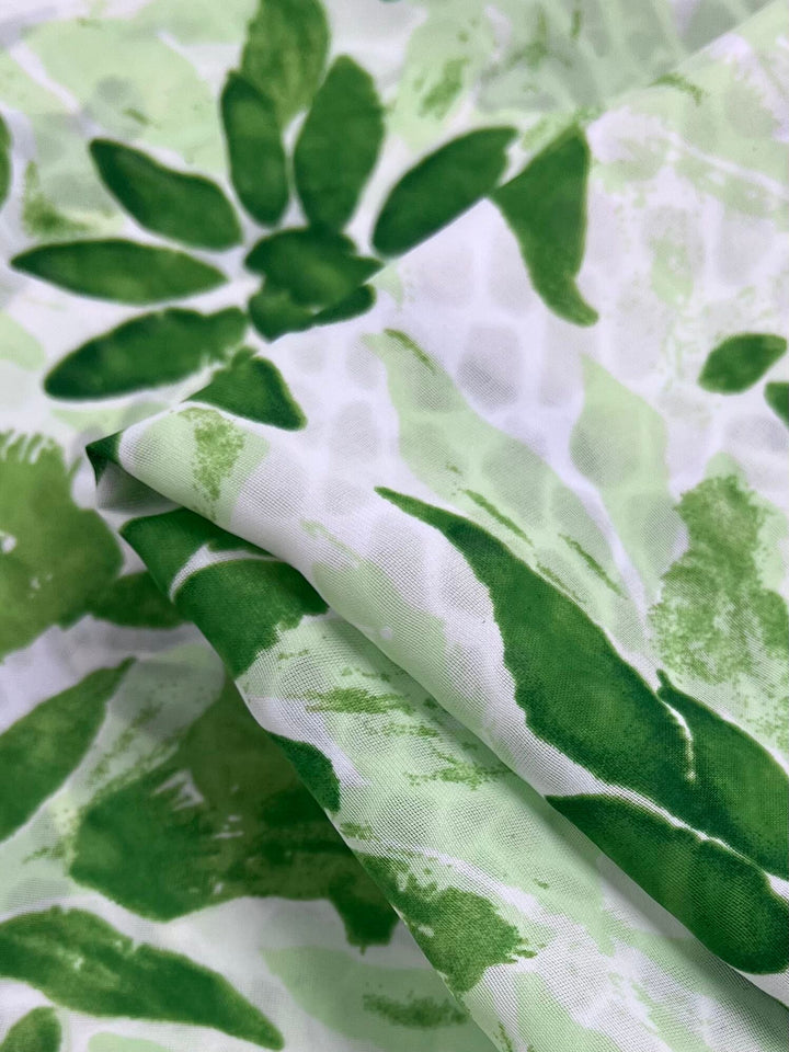 A close-up of Super Cheap Fabrics' "Salt Shrunk Print - Edelweiss - 155cm" reveals an elegant, green and white abstract leaf pattern. The fabric's folds showcase various green shades, creating a vibrant textured appearance that enlivens the print.