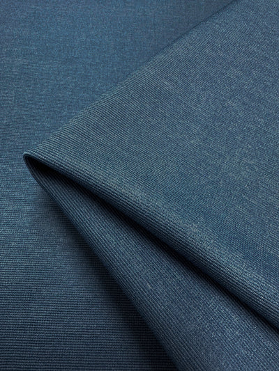 Here's a close-up of a folded piece of Ponte - Bluesteel - 157cm by Super Cheap Fabrics. This heavy-weight knit fabric boasts a soft, woven appearance with a subtle heathered pattern, featuring fine horizontal and vertical lines in its design. The fabric, in an elegant medium blue shade, is also wrinkle-resistant and appears neatly folded over itself.