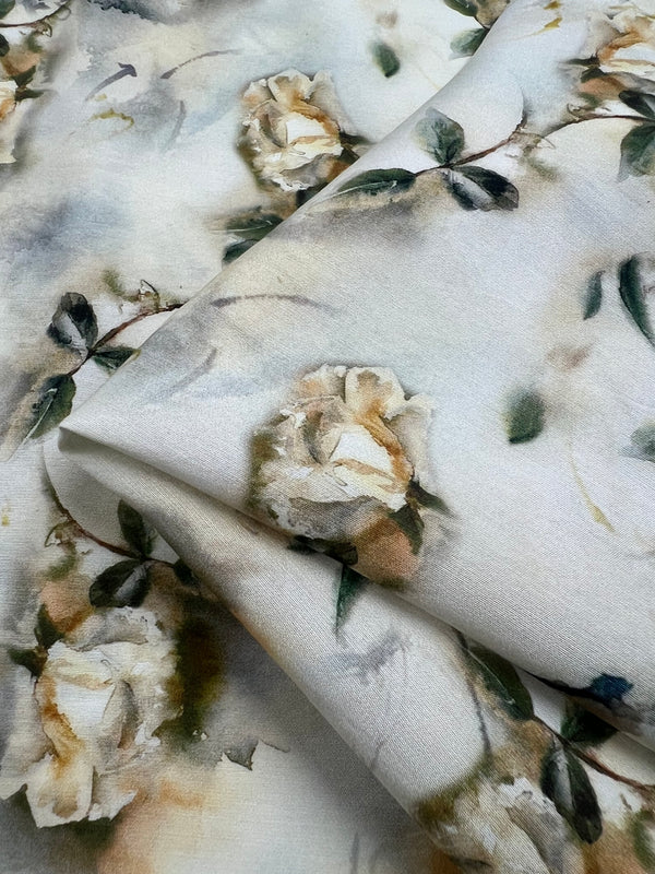 A close-up of a piece of light-weight fabric showcases a delicate floral pattern. The design features soft, white roses and green leaves against a light, almost watercolor-like background, lending a serene and elegant feel to the material—ideal for children's clothing. This is Designer Cotton - Painted Roses - 145cm from Super Cheap Fabrics.