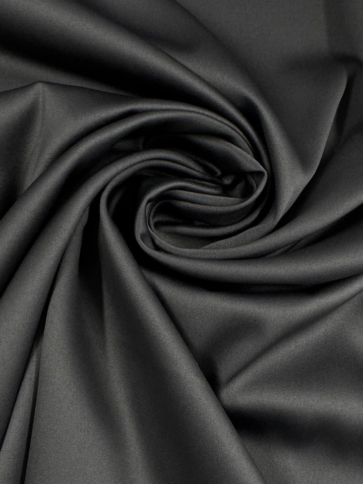 A close-up of Super Cheap Fabrics' Satin Deluxe in Charcoal (150cm) reveals its smooth, lightweight polyester satin twisted into a spiral, showcasing its soft, silky texture with subtle highlights and shadows.