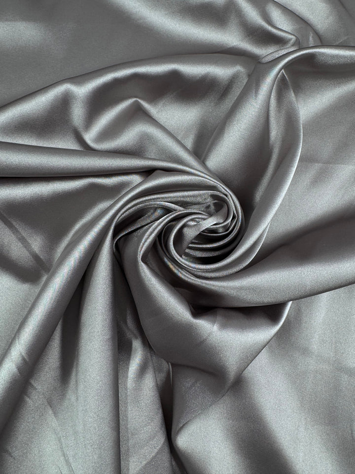 Close-up of Super Cheap Fabrics' Satin Deluxe in Satellite, 150cm, artfully twisted into a spiral. The glossy metallic gray satin reflects light softly off its folds, presenting a luxurious and elegant look.