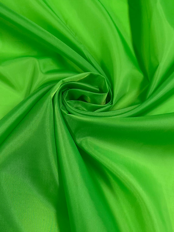 A close-up of a twisted piece of bright green fabric, showcasing its smooth texture and slight sheen. The lightweight lining fabric, "Lining - Green Flash - 150cm" by Super Cheap Fabrics, appears to be light and slightly translucent, with folds converging in a spiral pattern in the center.