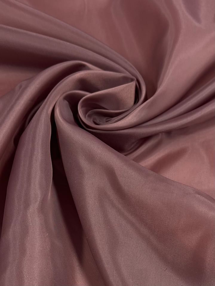 A close-up image of a piece of Super Cheap Fabrics' Lining - Burlwood - 150cm in a rich, dusty rose color. The lightweight fabric is smooth, with a slight sheen, and is elegantly gathered and folded into soft, swirling folds, creating a sense of texture and depth.