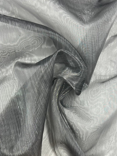 A close-up image of the Organza - Silver Filigree fabric from Super Cheap Fabrics, featuring a wavy, textured pattern in shades of grey and white. The material is lightly folded, highlighting its glossy, smooth surface and intricate weaves.