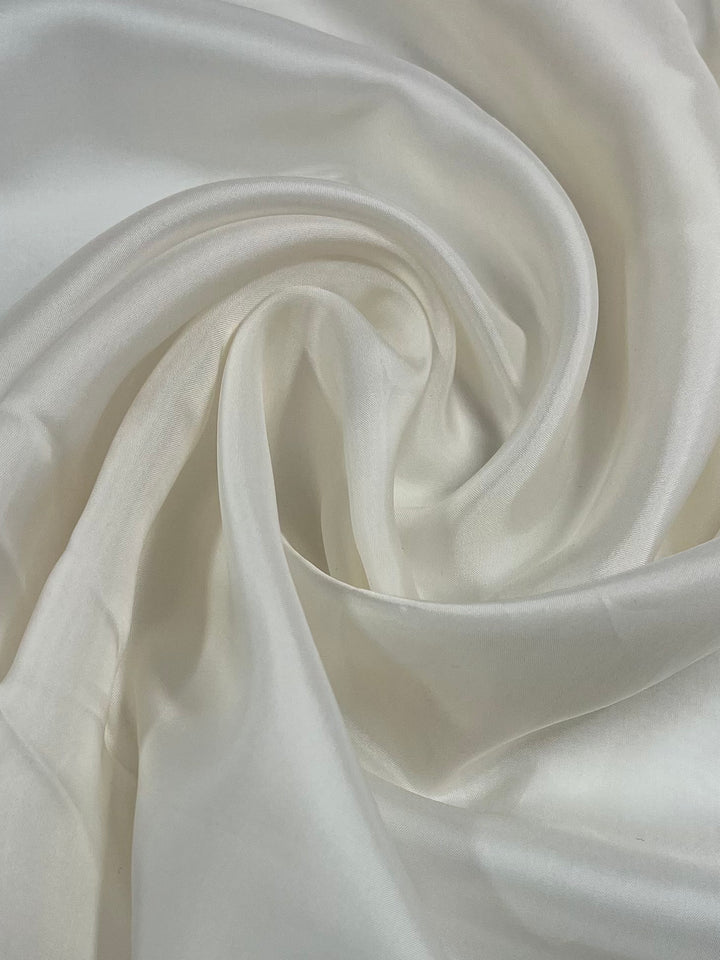 Close-up image of white silk fabric, intricately folded to create gentle waves and swirls. The delicate texture and soft sheen reflect light, highlighting the luxurious quality of the material, complemented by a lightweight 72 GSM polyester lining, Lining - Whisper White - 120cm from Super Cheap Fabrics.