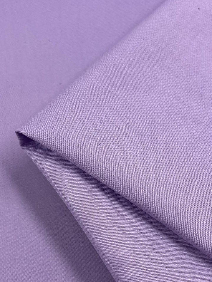 A close-up image of folded, delicate Cotton Drill fabric in a Regal Orchid hue from Super Cheap Fabrics, showcasing its smooth yet slightly textured surface. The lightweight Cotton Drill creates gentle shadows that highlight its soft drape and uniform color across the 110cm width.