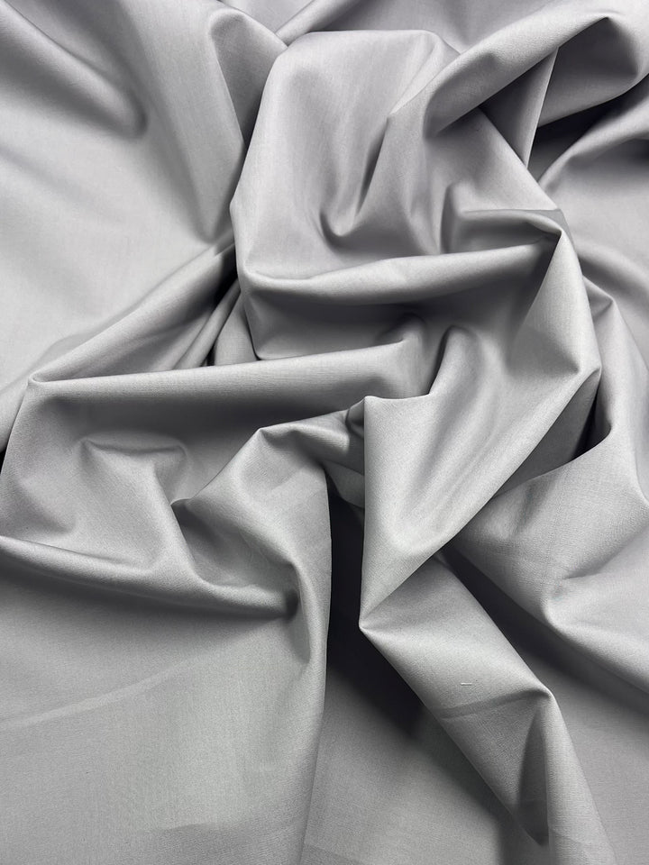 A close-up of the Super Cheap Fabrics' Plain Cotton in Grey, featuring smooth, soft folds and creases that showcase its texture and subtle sheen. The lightweight 150cm material appears slightly reflective, creating shadows and highlights on its surface.