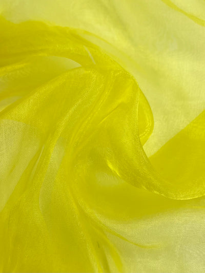 A close-up of the vibrant Quince yellow sheer organza fabric by Super Cheap Fabrics, softly draped and creating gentle waves and folds. This lightweight material allows light to produce subtle highlights and shadows on its surface, enhancing its delicate appearance. The fabric is available in a width of 150cm.