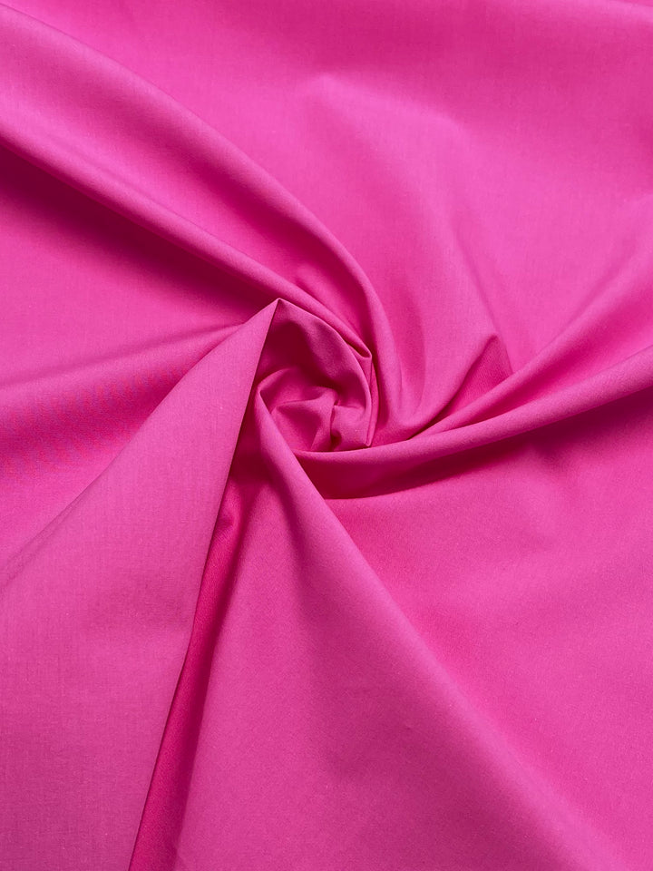 A detailed view of the Plain Cotton - Pink Power fabric from Super Cheap Fabrics reveals its vibrant bright pink shade, smoothly textured and carefully gathered to form a swirling pattern in the center. This lightweight material feels soft to the touch and its luminosity enhances its bold color, truly showcasing its pink power.