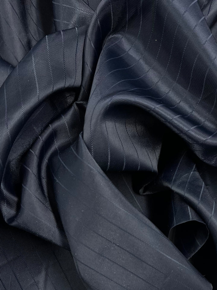 Close-up of crumpled, dark, pinstriped fabric. The material appears silky and has fine, diagonal lines running across its surface, creating a textured and elegant look suitable for refined tailoring. This is the Lining - Navy/ Stripe - 150cm from Super Cheap Fabrics.