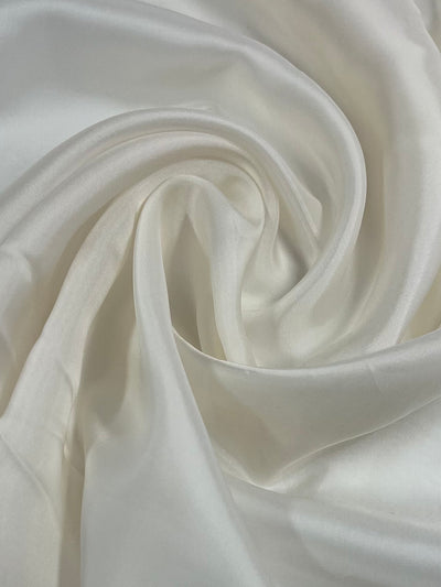 Close-up image of soft, white 72 GSM fabric draped and twisted into gentle folds and curves, creating a smooth and luxurious texture. The Super Cheap Fabrics Lining - Whisper White - 120cm has a subtle sheen, adding to its elegant appearance.