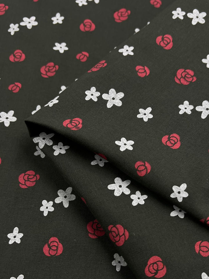 A piece of Linen Voile - Roses - 145cm from Super Cheap Fabrics, adorned with a floral pattern featuring small white flowers and red roses. The fabric is folded, showcasing the design prominently. The seamless repeating pattern adds a delicate, charming touch to the material, making it perfect for elegant home decor and offering excellent breathability.