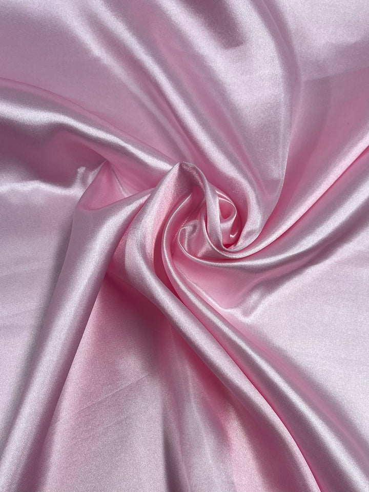 A close-up image of smooth, glossy Satin - Baby Pink - 150cm from Super Cheap Fabrics, showcasing its shiny surface and soft texture. The glamorous satin is artistically arranged in loose folds, creating gentle curves and swirls that highlight its luxurious quality.