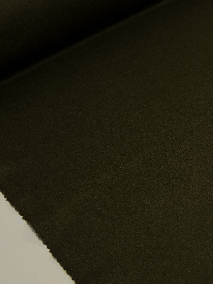 A close-up of a roll of Super Cheap Fabrics' "Wool Cashmere - Green Olive - 150cm," partially unrolled, revealing a slight texture on the dark olive green surface. The material looks smooth and tightly woven, ideal for crafting outer coats, with a straight edge visible at the bottom part of the image.