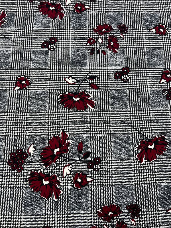 The Scuba Crepe - Hounds Dream from Super Cheap Fabrics, with a width of 160cm, features a striking black and white checkered houndstooth design adorned with scattered red and white flowers in various sizes. The vibrant flowers create a beautiful contrast against the detailed houndstooth background, all crafted from textured lightweight Scuba crepe fabric.