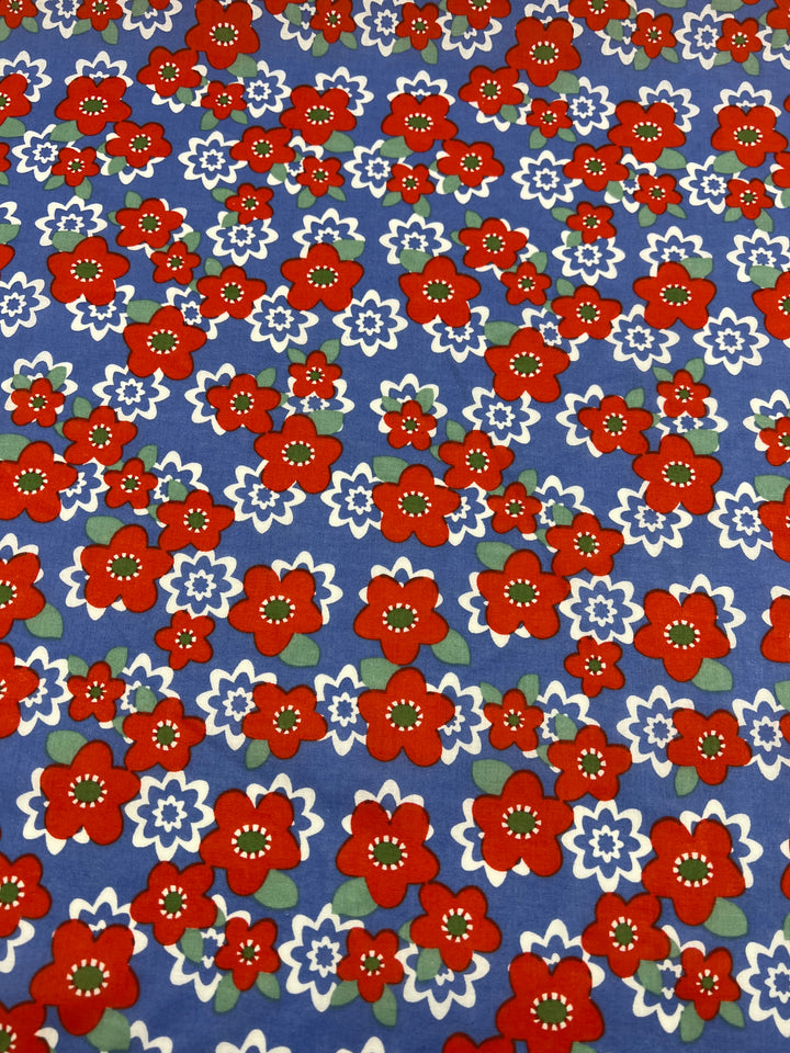 A vibrant multi-use fabric pattern featuring bold red and white flowers with green leaves on a blue background. Super Cheap Fabrics' Printed Cotton - Ramona - 150cm is made from 100% cotton, the lightweight fabric has flowers scattered across it, creating a lively and colorful design.