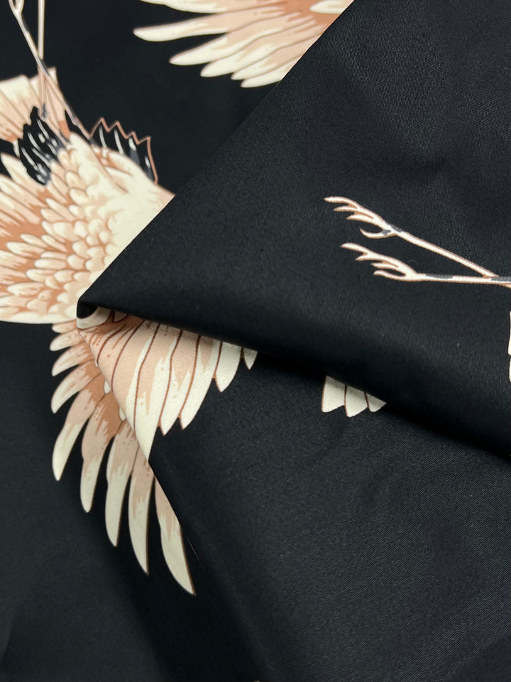 A close-up view of a Cotton Sateen fabric from Super Cheap Fabrics, named "Fly High - 130cm," showcases an intricate design of cream-colored birds on a dark background. The medium-weight material is folded to reveal detailed feathers and wing tips, with the solid black backdrop making this striking pattern ideal for clothing.