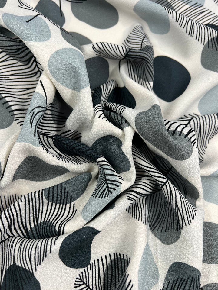 A close-up photo of Super Cheap Fabrics' Printed Crepe - Black Dotty Leafs, featuring a high-fashion, modern abstract pattern. The design showcases large, overlapping shapes in shades of grey and black with thin, wavy lines resembling leaves or feathers on a white background. This lightweight fabric is slightly gathered and measures 150cm across.