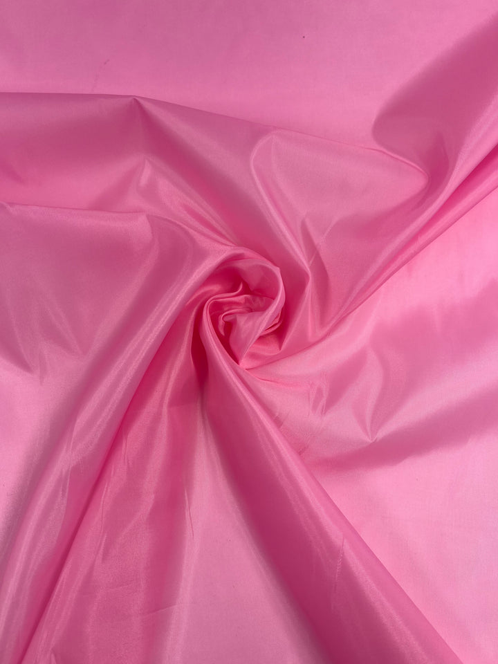 A close-up image of a piece of bright pink, glossy fabric with a smooth texture. The vibrant *Super Cheap Fabrics* Lining - Pink - 150cm is slightly gathered and twisted at the center, creating subtle wrinkles and folds around the area. The lighting enhances the fabric’s sheen and vibrant color.