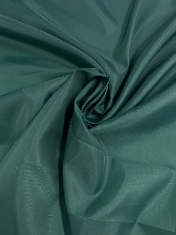 A close-up of Super Cheap Fabrics' Lining - Evergreen - 150cm, a smooth, silky polyester lining in a deep emerald green color. The lightweight lining fabric is slightly gathered and twisted in the center, creating soft folds and a subtle shine that reflects the light.