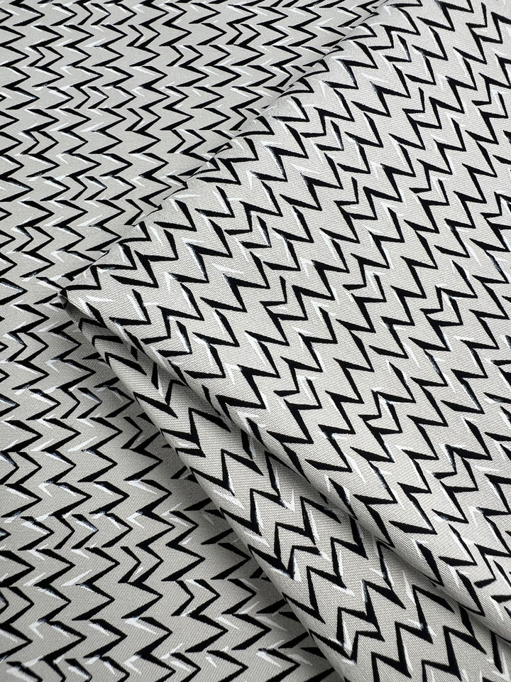 Close-up of folded Stretch Bengaline - Vibration - 150cm from Super Cheap Fabrics, featuring a striking black and white zigzag pattern. The sharp, consistent zigzags create a bold geometric design across this two-way stretch medium-weight fabric. The smooth texture appears slightly reflective under the light.