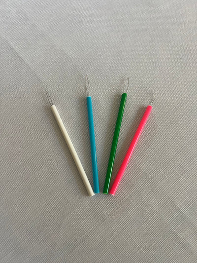 Four Super Cheap Fabrics Needle Threaders are arranged on a light gray textured background. The threaders have thin metal loops at one end and colorful plastic handles in white, blue, green, and pink.