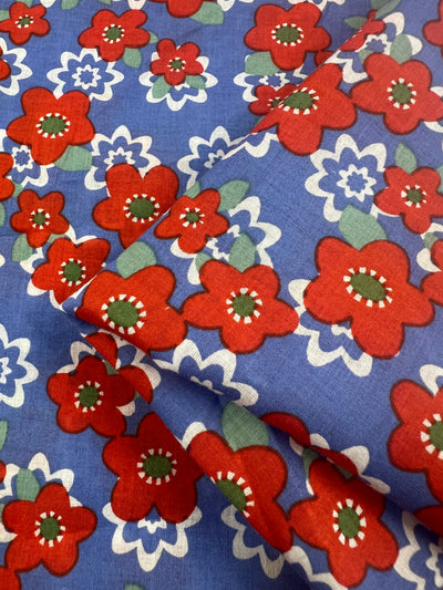 A close-up view of the Printed Cotton - Ramona - 150cm by Super Cheap Fabrics featuring a colorful floral pattern. The blue background is adorned with large red flowers with green centers, white petals, and smaller white and blue flowers interspersed throughout. The lightweight fabric is slightly folded, creating a textured look perfect for multi-use purposes.