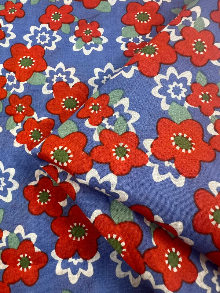 A close-up view of the Printed Cotton - Ramona - 150cm by Super Cheap Fabrics featuring a colorful floral pattern. The blue background is adorned with large red flowers with green centers, white petals, and smaller white and blue flowers interspersed throughout. The lightweight fabric is slightly folded, creating a textured look perfect for multi-use purposes.