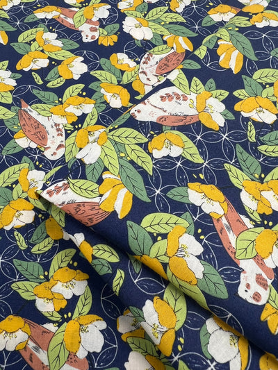 A close-up of a fabric design featuring an intricate botanical print of yellow flowers, green leaves, and beige mushrooms with orange caps on a dark navy blue background. This Printed Cotton - Wild Camellias - 150cm by Super Cheap Fabrics appears neatly folded in layers, perfect for multiple uses.