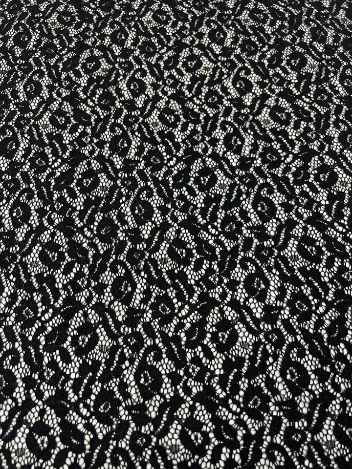 A close-up image of a lightweight Super Cheap Fabrics Black Lace - Ora - 150cm with an intricate and delicate pattern, creating a textured appearance. The design consists of various swirls and loops, with the lace's holes forming an almost floral-like arrangement against a contrasting background.