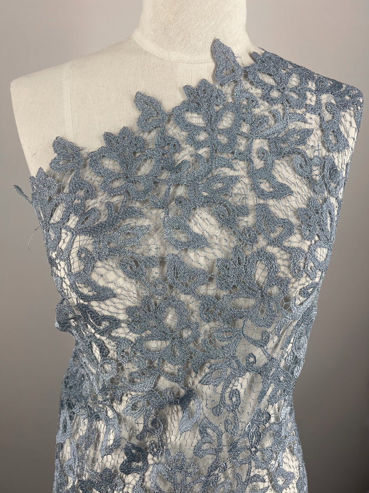 A dress form is draped in intricate Evening Lace - Metallic Blue - 120cm by Super Cheap Fabrics with a floral pattern. The lace is arranged asymmetrically over one shoulder, revealing a sheer mesh underneath. The background is a plain off-white wall.