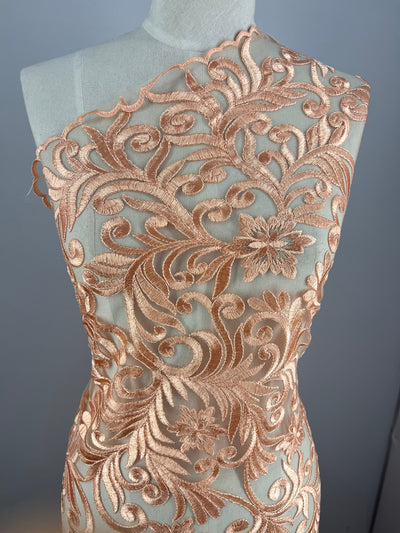 A dressmaker's mannequin is draped with an intricate fabric featuring copper-toned, ornate floral and leaf embroidery, creating a swirling design. The semi-sheer Evening Lace - Galadriel from Super Cheap Fabrics covers the upper body diagonally from the left shoulder to the right hip.