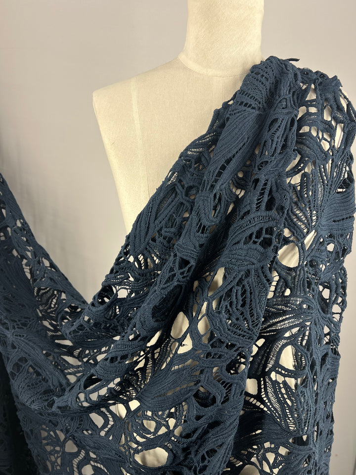 A mannequin is draped with an intricate, dark teal crochet lace fabric crafted from lightweight polyester, featuring an elaborate, openwork design that showcases various patterns and textures. The backdrop is a simple gray setting. The fabric used is Evening Lace - Navy - 100cm from Super Cheap Fabrics.