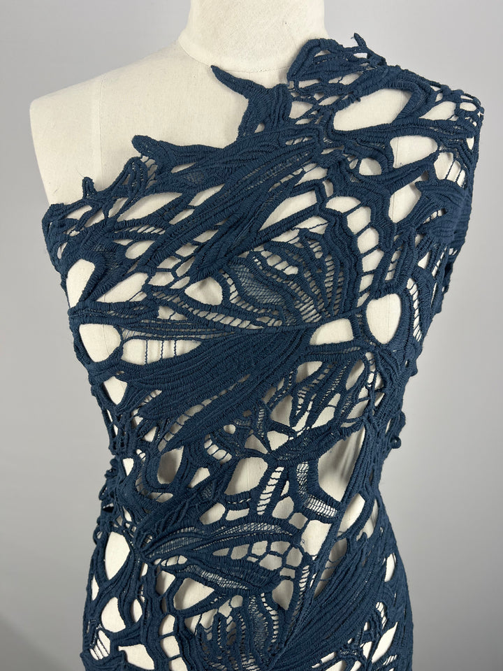 A dress form displaying an Evening Lace - Navy - 100cm garment made of intricately woven, navy blue yarn from Super Cheap Fabrics. The design features abstract, organic patterns with large cut-out sections, giving it a lace-like appearance similar to navy lace. The background is plain and light gray.