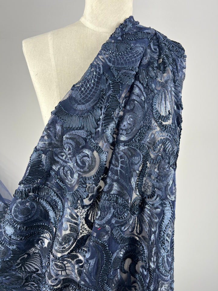 A close-up of a draped, intricately patterned navy blue, 100% polyester fabric with embroidered floral and paisley designs displayed on a white mannequin. The medium weight fabric and detailed embroidery create a textured, elegant appearance. This is Evening Lace - Night Shade - 150cm by Super Cheap Fabrics.