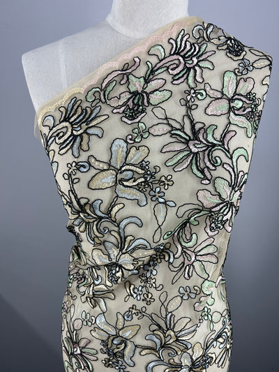 An Embroidered Lace - Yasmin - 150cm by Super Cheap Fabrics adorned with intricate floral embroidery in various colors is displayed draped over a white mannequin. The design includes flowers and leaves on a sheer, neutral-colored background, creating an elegant and detailed pattern.