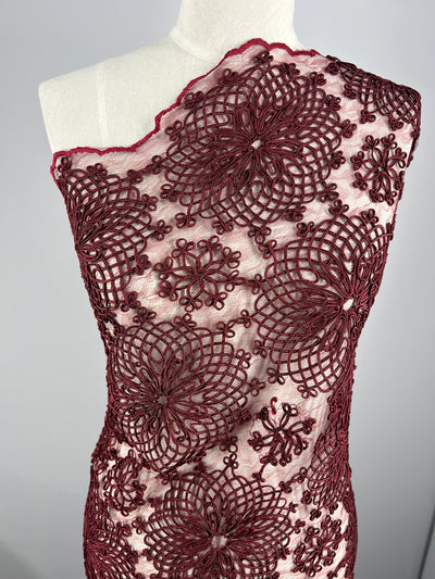 A close-up of a dress with an Evening Lace - Elizabeth - overlay in detailed burgundy lace, featuring intricate floral patterns. The lace sits on a sheer, light-colored fabric background, draped over a dress form. The dress has a one-shoulder design by Super Cheap Fabrics.