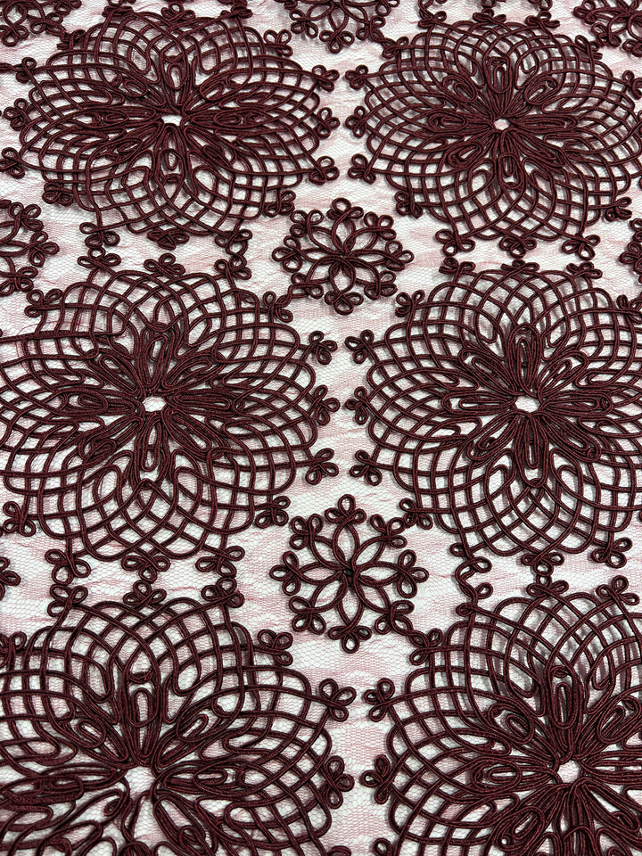 The intricate Evening Lace - Elizabeth - features repeating floral patterns in a rich burgundy color against a light background. The design includes large flowers with interconnected loops and detailed centers, creating a delicate and elegant texture. This exquisite fabric is offered by Super Cheap Fabrics.