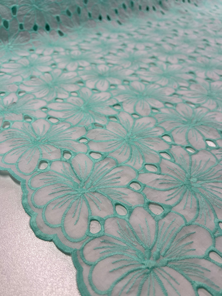 Close-up of a light teal Broderie Anglaise - Daised Pastel - 125cm fabric from Super Cheap Fabrics showcasing intricate, floral lace patterns. The design features repeating blossoms with delicate details, and the fabric has a scalloped edge, enhancing its elegance. The background is softly blurred.