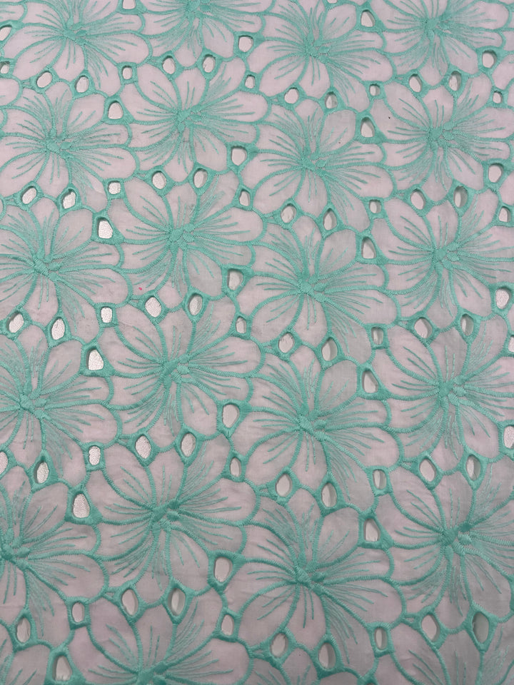 A close-up of a fabric with an intricate floral pattern. The Super Cheap Fabrics Broderie Anglaise - Daised Pastel - 125cm features large, light green flowers with detailed petals and leaves against a soft pink background. The flowers are evenly spaced, creating a delicate, lace-like design with small cutouts.