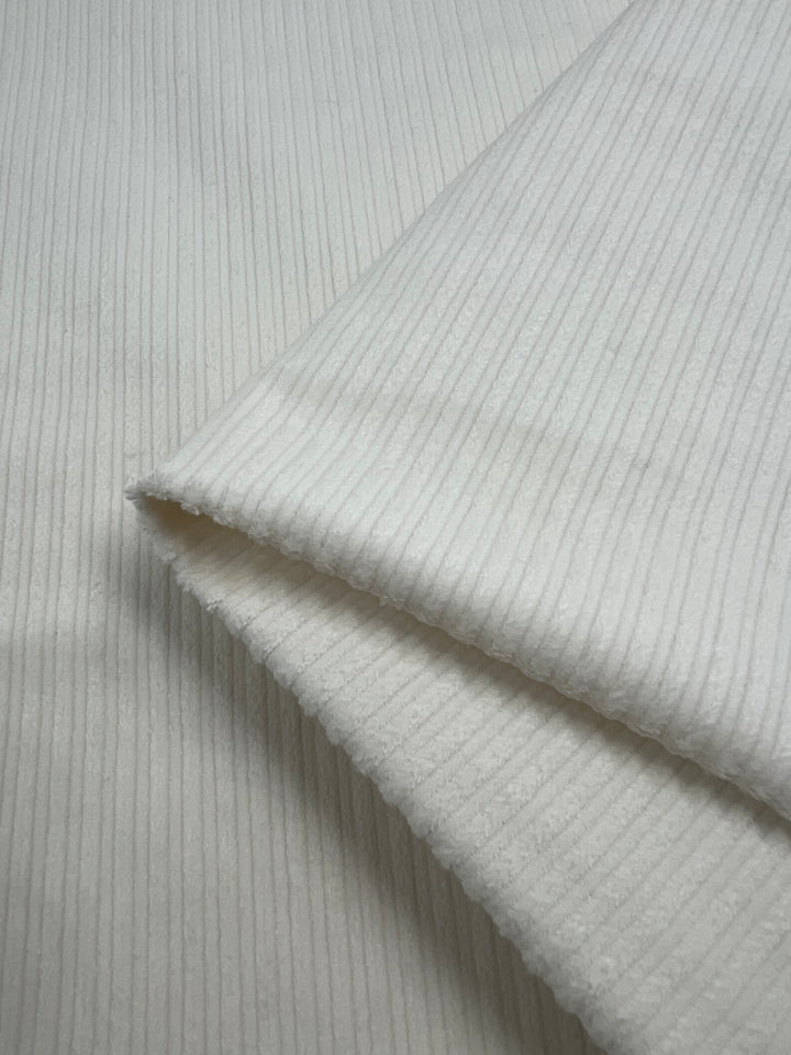 Close-up of a folded piece of Medium Wale Stretch Corduroy – Ivory – 150cm by Super Cheap Fabrics. The ribbed fabric features uniform, narrow vertical stripes with a slight sheen. Its soft and slightly plush texture suggests that it would be ideal for cozy clothing or upholstery, thanks to its medium to heavy weight.