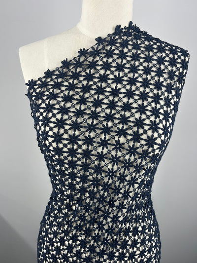 A mannequin is dressed in a fitted garment featuring a navy floral crochet pattern made from Super Cheap Fabrics' Anglaise Lace. The design forms a series of interconnected flowers that cover the entirety of the dress, creating a stylish and intricate texture. The dress has an asymmetrical, one-shoulder neckline.