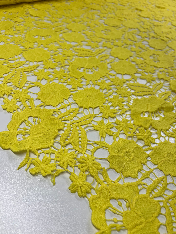 A close-up view of Super Cheap Fabrics' Anglaise Lace in bright yellow, featuring an intricate floral pattern. The 127cm wide lace is laid out on a flat surface, showcasing the detailed textures and openwork design of the material—perfect for bridal wear or elegant home décor projects.