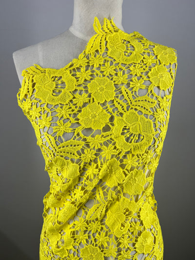 A mannequin showcases a vibrant yellow, intricately crocheted one-shoulder dress made from Super Cheap Fabrics' Anglaise Lace (127cm), adorned with large floral patterns. The asymmetrical design exposes one shoulder and highlights detailed craftsmanship akin to fine lace fabrics, set against a neutral gray background.