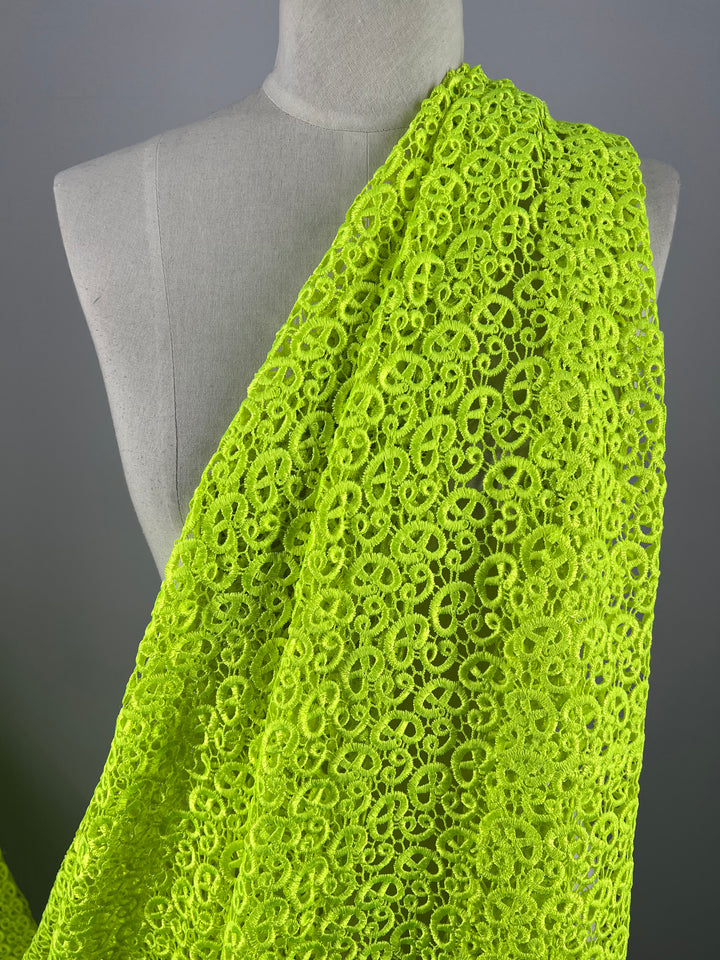 A close-up image showcases the Anglaise Lace fabric in Sharp Green draped over a gray dress form. This intricate lace from Super Cheap Fabrics features a repeating circular pattern, creating a textured and vibrant appearance against the neutral background of the mannequin. It's perfect for bridal wear or home décor projects.